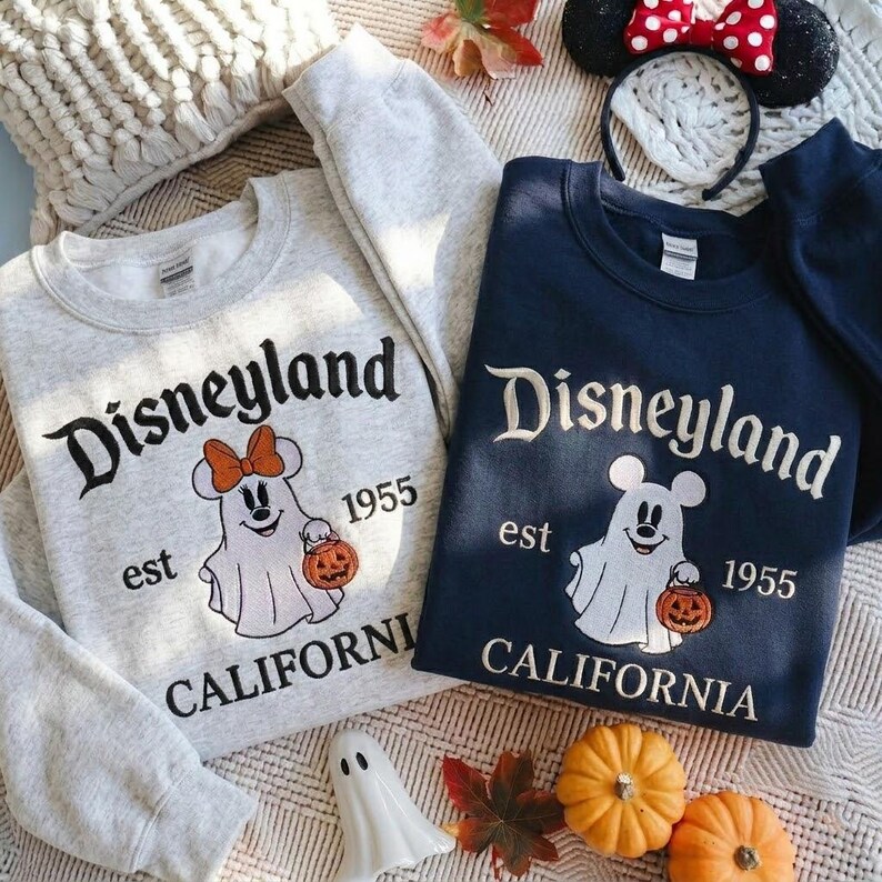 May include: Two crewneck sweatshirts, one light gray and one navy blue, featuring the text Disneyland est 1955 California. Each displays a white ghost character wearing mouse ears and holding a jack-o'-lantern pumpkin, with an orange bow on the gray.