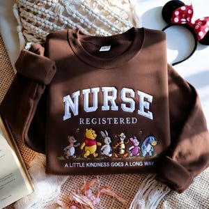 May include: A brown sweatshirt with the word "NURSE" in white, embroidered letters. Below, the word "REGISTERED" is embroidered. A line of cartoon characters is embroidered below the text, with the phrase "A LITTLE KINDNESS GOES A LONG WAY".