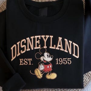 May include: Black sweatshirt with tan embroidered "DISNEYLAND" text and a Mickey Mouse graphic. Below the text is "EST. 1955". The sweatshirt has a crew neck and long sleeves. The design is reminiscent of vintage theme park apparel.