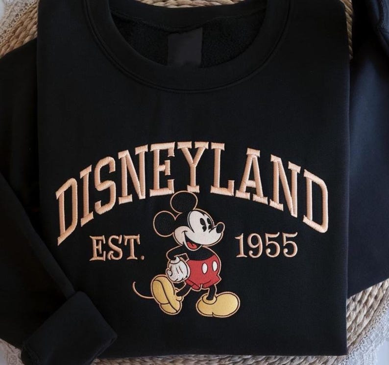 May include: Black sweatshirt with tan embroidered "DISNEYLAND" text and a Mickey Mouse graphic. Below the text is "EST. 1955". The sweatshirt has a crew neck and long sleeves. The design is reminiscent of vintage theme park apparel.