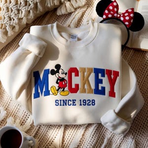 May include: Cream-coloured sweatshirt with the word "Mickey" in blue, red, and gold lettering, and a cartoon Mickey Mouse graphic. The text "SINCE 1928" is embroidered below. A Minnie Mouse ear headband with a red and white polka dot bow is visible.