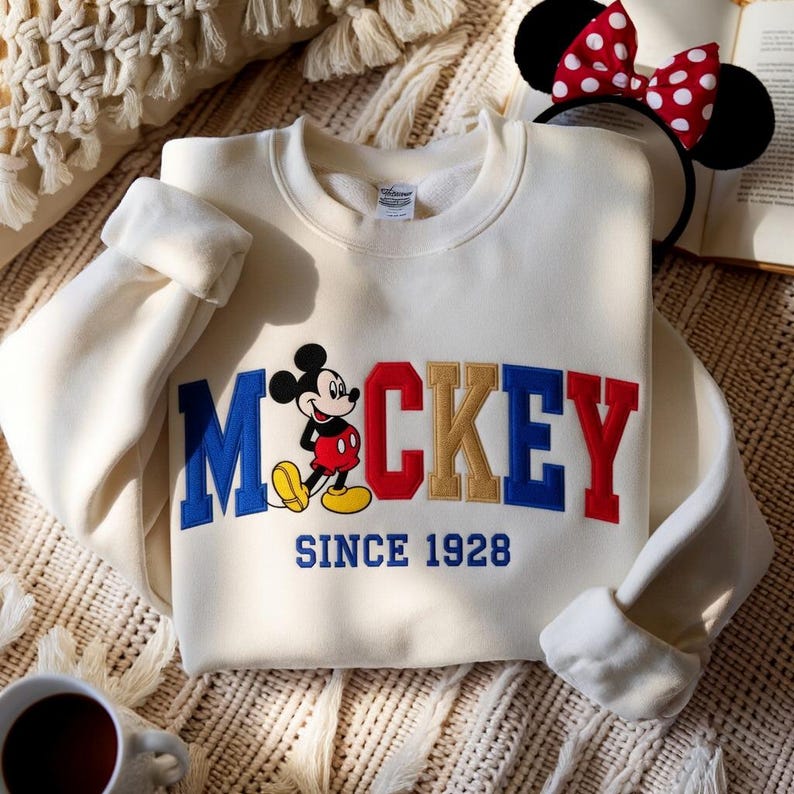 May include: Cream-coloured sweatshirt with the word "Mickey" in blue, red, and gold lettering, and a cartoon Mickey Mouse graphic. The text "SINCE 1928" is embroidered below. A Minnie Mouse ear headband with a red and white polka dot bow is visible.