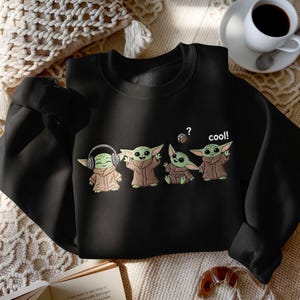 May include: Black sweatshirt featuring four Grogu illustrations. One Grogu wears headphones, another has a question mark above its head, and the last one says "cool!". The sweatshirt is made of a soft material.