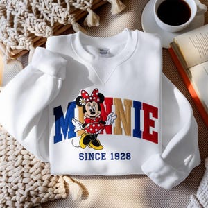 K&ouml;nnte beinhalten: Wei&szlig;es Sweatshirt mit einem Minnie Mouse-Motiv. Der Schriftzug "MINNIE" ist in Rot, Gold und Blau dargestellt, mit Minnie Mouse in einem roten gepunkteten Kleid. Der Text "SINCE 1928" steht darunter.