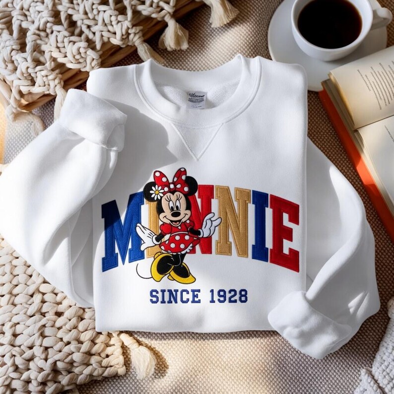 K&ouml;nnte beinhalten: Wei&szlig;es Sweatshirt mit einem Minnie Mouse-Motiv. Der Schriftzug "MINNIE" ist in Rot, Gold und Blau dargestellt, mit Minnie Mouse in einem roten gepunkteten Kleid. Der Text "SINCE 1928" steht darunter.