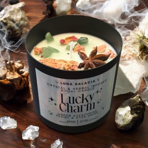 Pode incluir: Uma vela preta com um rótulo que diz "Lucky Charm". A vela tem cera verde clara, adornada com cristais verdes, flores secas e anis estrelado. É uma "Crystal & Herbal Infused Ritual Candle".
