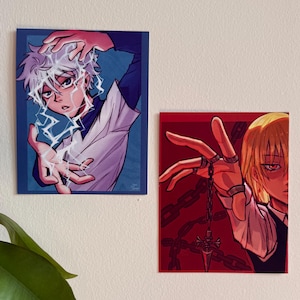 May include: Two rectangular art prints featuring anime-style characters. The left print shows a white-haired character with blue lightning effects. The right print depicts a blonde character with red eyes holding a chain with a pointed pendant.