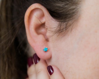 May include: A close-up of a person's ear with a small turquoise stud earring. The earring is set in gold and has a simple, elegant design.