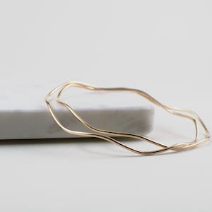 May include: Two delicate, gold-colored bangles with a wavy design. The bracelets are displayed on a white marble surface, creating a minimalist aesthetic. The thin, flowing lines of the jewelry suggest a modern, elegant style.