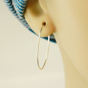 May include: A close-up of a silver hoop earring. The earring is thin and oval-shaped, with a smooth, reflective surface. The earring is in the ear of a person with fair skin. A blue and white patterned fabric is visible in the background.