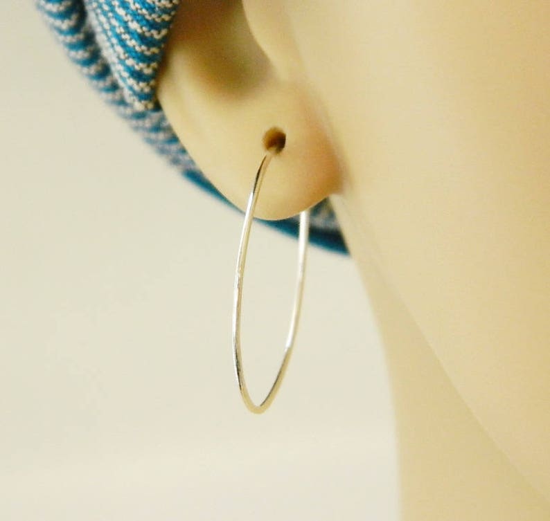 May include: A close-up of a silver hoop earring. The earring is thin and oval-shaped, with a smooth, reflective surface. The earring is in the ear of a person with fair skin. A blue and white patterned fabric is visible in the background.