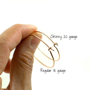May include: Two copper-colored hoop earrings are held in a hand against a white background. One earring is labeled "Skinny 20 gauge" and the other is labeled "Regular 18 gauge". The earrings are simple and elegant.
