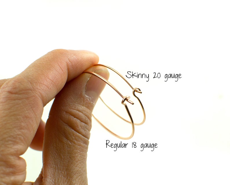 May include: Two copper-colored hoop earrings are held in a hand against a white background. One earring is labeled "Skinny 20 gauge" and the other is labeled "Regular 18 gauge". The earrings are simple and elegant.
