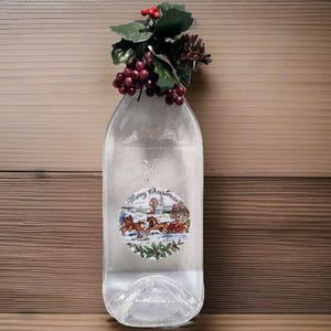 以下が含まれることがあります： 「メリークリスマス」と馬車を描いた白いラベルが貼られた透明なガラス瓶。赤い実と緑の葉のガーランドで飾られています。