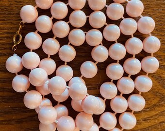 May include: A vintage necklace featuring numerous light pink, round beads strung together. The beads are evenly spaced and connected by a thin, white string. The necklace has a gold-colored clasp. The background is a wooden surface.