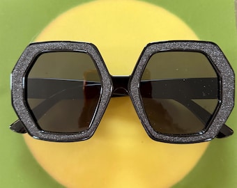 Peut inclure: Lunettes de soleil hexagonales dotées d'une monture noire avec une finition texturée et pailletée, accompagnées de verres teintés foncés. Les montures géométriques présentent une forme à six côtés, offrant un aspect audacieux et uniforme.
