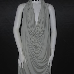 May include: A light gray, floor-length dress with a draped cowl neckline. The dress is displayed on a white mannequin against a black background. The mannequin's neck has the letters "HEF" printed on it.