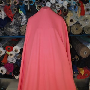 May include: A large piece of coral pink fabric draped over a stand. The fabric is smooth and appears to be a lightweight material. The fabric is surrounded by rolls of fabric in various colors and patterns.