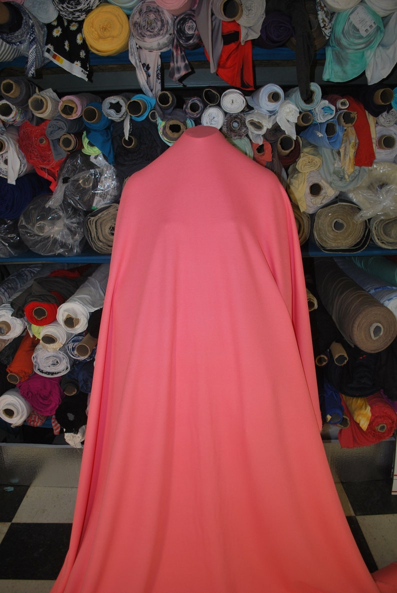 May include: A large piece of coral pink fabric draped over a stand. The fabric is smooth and appears to be a lightweight material. The fabric is surrounded by rolls of fabric in various colors and patterns.