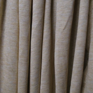 Puede incluir: Primer plano de una tela de punto de canal&eacute; beige con una textura sutil. La tela est&aacute; doblada y drapeada, creando un aspecto suave y fluido.