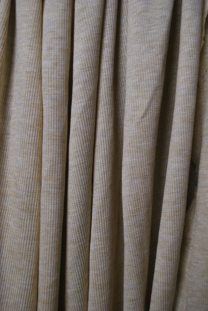 Puede incluir: Primer plano de una tela de punto de canal&eacute; beige con una textura sutil. La tela est&aacute; doblada y drapeada, creando un aspecto suave y fluido.