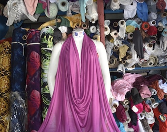 May include: A mannequin wearing a fuchsia-colored draped fabric garment. The mannequin is standing in front of a wall of fabric rolls in various colors and patterns.