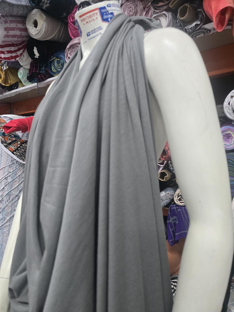 Op de afbeelding: Een effen grijze stof gedrapeerd over een witte etalagepop-torso. De stof heeft een gladde, matte textuur en is geplooid bij de halslijn, waardoor verticale plooien ontstaan die naar beneden vallen. De nek van de etalagepop toont een klein Priority Mail-label.