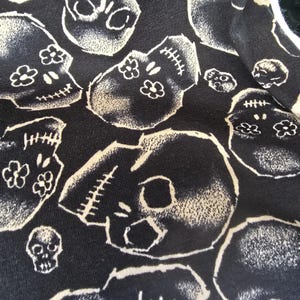 May include: Black fabric with a repeating pattern of white skull illustrations. The skulls have stitched details and floral accents. The design is a dark, gothic style.