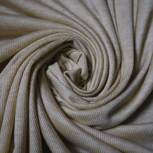 Puede incluir: Primer plano de una tela de punto de canal&eacute; beige, retorcida en forma de espiral. La tela tiene un brillo sutil y una superficie suave y texturizada.