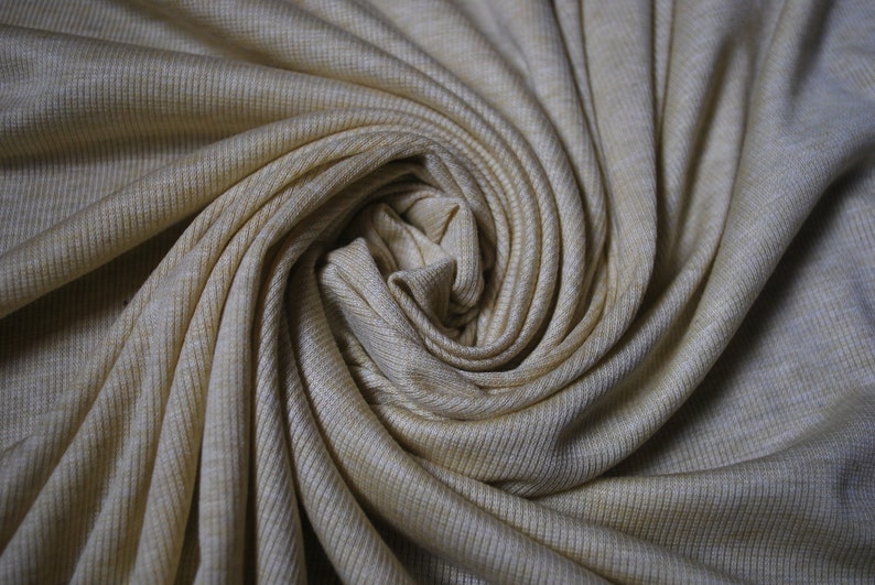 Puede incluir: Primer plano de una tela de punto de canal&eacute; beige, retorcida en forma de espiral. La tela tiene un brillo sutil y una superficie suave y texturizada.