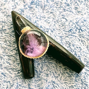 Puede incluir: Un colgante con una piedra de amatista redonda y transparente, con inclusiones moradas, engastada en un marco de cuerda beige trenzada. Descansa sobre un objeto de madera marrón oscuro con un extremo puntiagudo. El fondo es una tela azul claro texturizada.