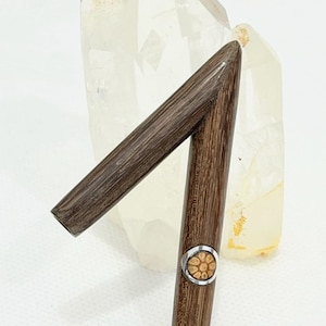 Puede incluir: Un portacigarros de madera marrón oscuro con una banda metálica decorativa y un diseño estampado. El soporte está en ángulo, sobre un fondo de cristal de cuarzo transparente.