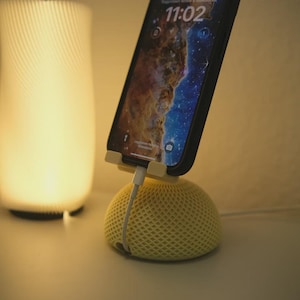 Könnte beinhalten: Ein gelber Telefonständer mit gewebtem Design hält ein Smartphone, das ein Galaxienbild anzeigt. Das Telefon wird mit einem weißen Kabel aufgeladen. Im Hintergrund befindet sich eine leuchtende weiße Lampe.