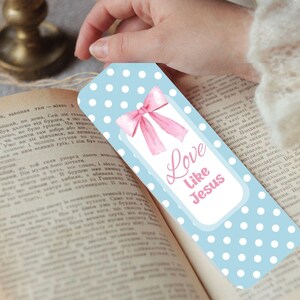 May include: A light blue bookmark with white polka dots and a pink bow illustration. The bookmark has the text "Love Like Jesus" in pink. The bookmark is held over an open book.