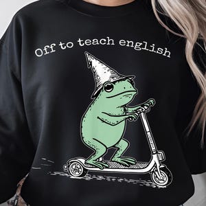May include: Black sweatshirt with the text "Off to teach english." The graphic shows a green frog wearing a wizard hat and riding a scooter. The frog is drawn in a cartoon style.