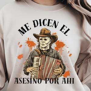 Könnte beinhalten: Beiges Sweatshirt mit einer Grafik einer maskierten Figur mit Cowboyhut, die Akkordeon spielt, mit orangefarbenen Spritzern. Der Text "ME DICEN EL ASESINO POR AHI" ist oberhalb und unterhalb der Figur aufgedruckt.
