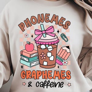 Puede incluir: Sudadera beige con un diseño gráfico colorido. El diseño incluye las palabras "PHONEMES" y "GRAPHEMES & caffeine" en letras naranjas y marrones. El gráfico también presenta una bebida de café, libros, un lápiz y una manzana.