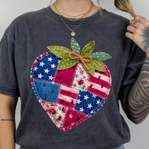 Puede incluir: Camiseta gris oscuro con un diseño de fresa de patchwork. La fresa está compuesta por parches de tela rojos, blancos y azules, incluyendo estrellas y rayas. La camiseta tiene cuello redondo y mangas cortas. La palabra "PEPPER" está impresa en la parte inferior.