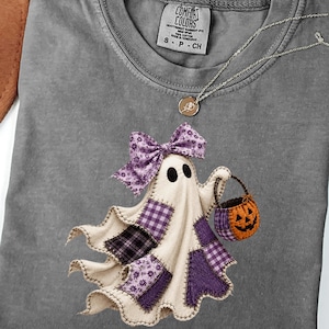 Può includere: Una t-shirt grigio antracite con la grafica di un fantasma decorato con toppe a quadri e motivi floreali viola, un grande fiocco viola sulla testa e un piccolo secchiello a forma di zucca arancione. La t-shirt ha un girocollo.