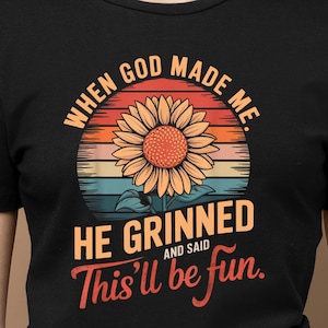 May include: Black t-shirt featuring a sunflower design with the text "When God Made Me, He Grinned and Said This'll be fun." The sunflower has orange and yellow petals with a brown center, set against a sunset background.