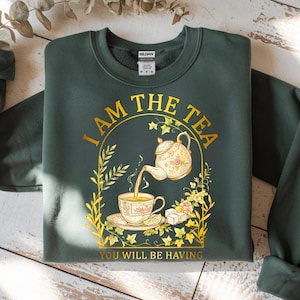 Può includere: Felpa verde scuro con la scritta "I AM THE TEA YOU WILL BE HAVING" in oro. Il design presenta una teiera che versa tè in una tazza, con dettagli floreali.
