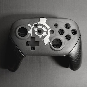 Peut inclure: Une manette de jeu vidéo noire et grise avec un motif graphique blanc sur le dessus. La manette comporte une croix directionnelle, plusieurs boutons et deux joysticks analogiques. Les poignées sont texturées.