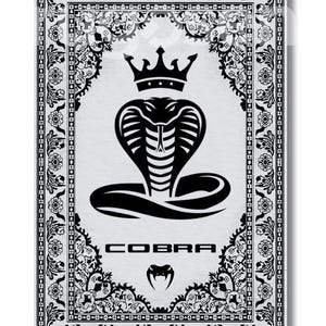 Op de afbeelding: Zwart-witte afbeelding met een cobra met een kroon, geplaatst in een sierlijke bloemenrand. Het woord "COBRA" staat onder de slangenafbeelding, met een kleiner cobra-embleem eronder. Het ontwerp staat op een witte achtergrond.