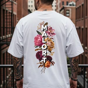 May include: White t-shirt featuring a floral design and the word "MALORT" in white block letters. The floral arrangement includes pink, yellow, and burgundy flowers. The shirt has a relaxed, oversized fit.