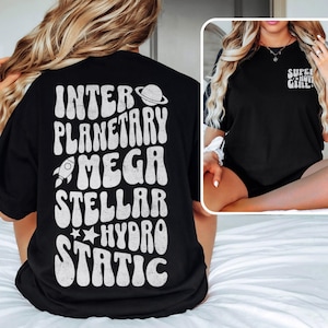 Puede incluir: Camiseta negra con texto blanco en la espalda: "INTERPLANETARY MEGA STELLAR HYDRO STATIC" con un cohete, un planeta y estrellas. La parte delantera de la camiseta tiene el texto "SUPER NOVA GIRL".