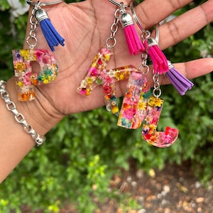 May include: A collection of keychains featuring the letters P, A, T, and J. Each letter is filled with colorful dried flowers and encased in clear resin. The keychains have silver rings and are adorned with blue, pink, and purple tassels.