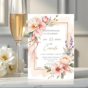 Op de afbeelding: Een uitnodiging met bloemaccenten en gouden tekst, inclusief de woorden "Acompáñanos a celebrar los 53 años de Camila." Een champagneglas met een drankje en witte bloemen staan op de voorgrond.