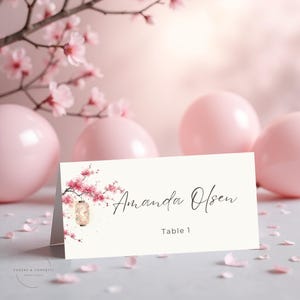 May include: A cream-coloured place card with the name "Amanda Olsen" in elegant script. The card features a watercolour illustration of pink cherry blossoms and a lantern. "Table 1" is printed below the name.