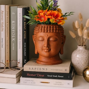 Puede incluir: Un jarrón de cabeza de Buda de terracota lleno de flores naranjas y moradas. El jarrón está sobre una estantería con libros, incluyendo "Sapiens" y "Homo Deus". También se ven otros libros y un jarrón decorativo.