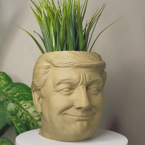 May include: A beige ceramic head-shaped planter with a smiling expression and closed eyes, filled with green artificial grass. The planter sits on a white, round base. The face has detailed features, including wrinkles and sculpted hair.
